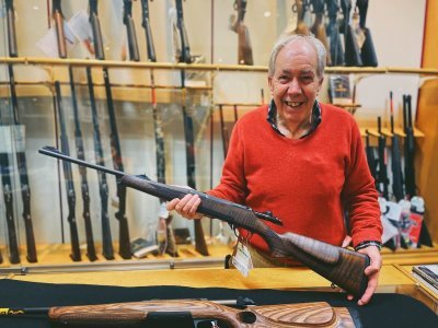 Waffenhändler Willi Becker rotes Oberteil hält eine Waffe vor Waffenschrank